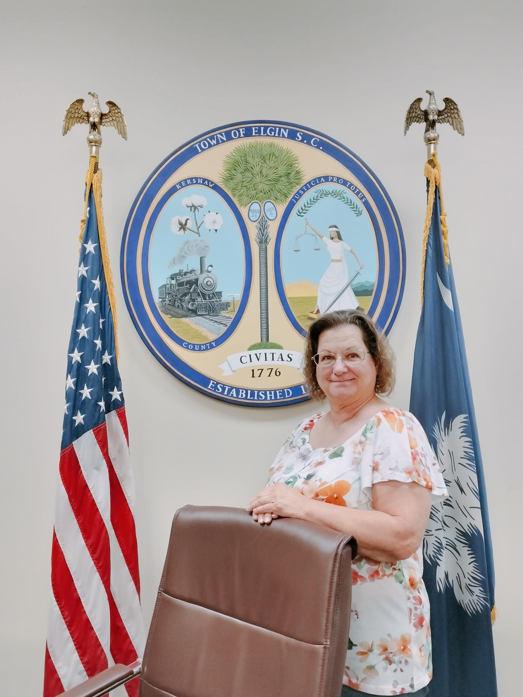 Woman standing by a chair in front of the Town of Elgin, SC seal, with US and state flags on either side, at the elgin municipal court.