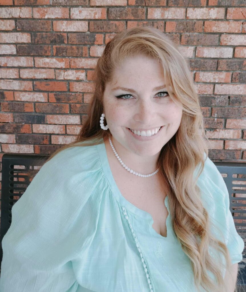 Smiling woman with long red hair in a light blue top and pearl jewelry, sitting in front of a brick wall near Elgin Town Hall.