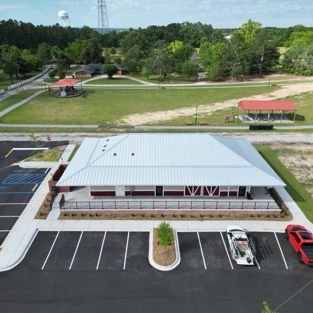 Aerial view of a modern building with a metal roof, parking lot, and green park area with trees and gazebos.